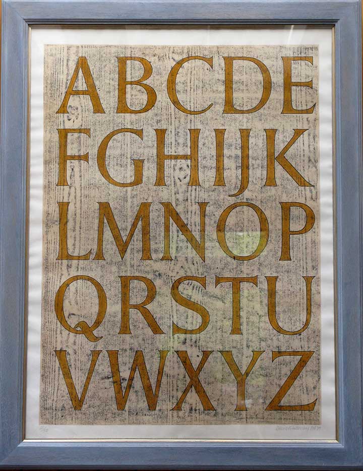 A hand written alphabet. The letters are all uppercase, in gold on a wood grain background