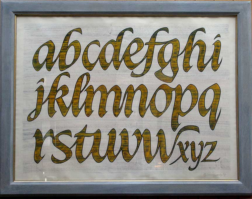a hand written alphabet in a frame. The letters are all lower case, in gold on a white background.