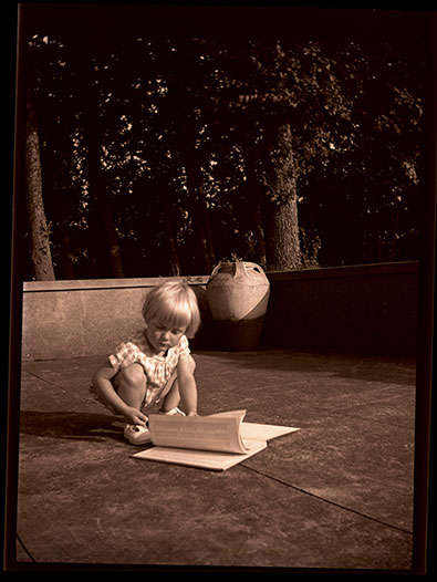 Black and white photo of a young boy reading a large book on a patio garden.
