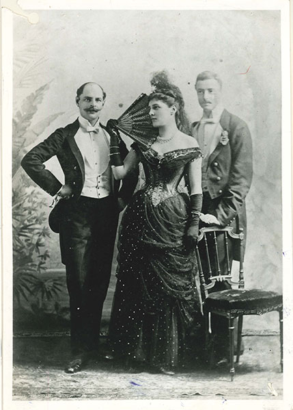 Black and white photograph of Lady Randolph Churchill standing with two men. All are in evening dress and Lady Randolph Churchill is holding a fan.