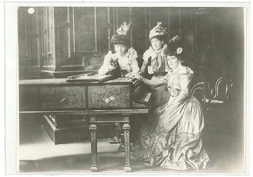 Black and white photograph of Lady Randolph Churchill seated at a piano with two female friends leaning on the top of the piano, where a black and white cat is lying.