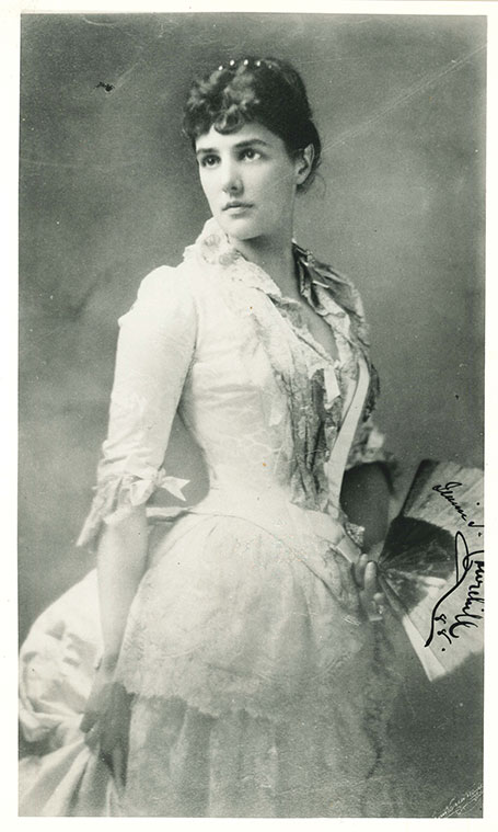Black and white portrait photograph of Lady Randolph Churchill. She is wearing a pale coloured dress and is holding a fan.