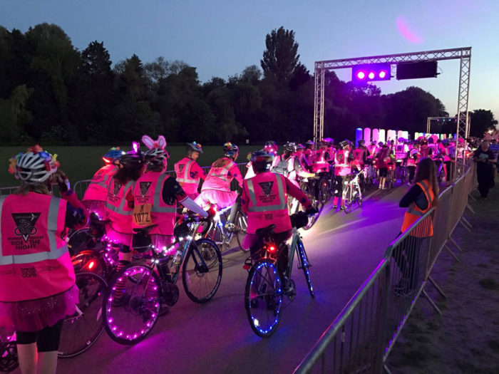 A large group of cyclists preparing to set off. The road has been lit up in pink light, everyone is wearing a pink high vis jacket, and many are wearing decorations such as bunny ears on their helmets. Some of the bicycles have been decorated as well with extra lights on their wheels and frames.