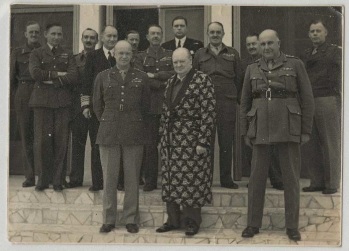 Black and white photo of Churchill in his dressing gown and slippers, posing for a photo with a group of men in military uniform
