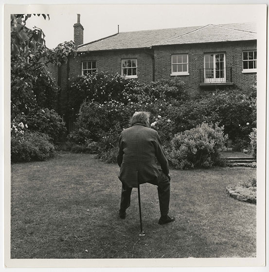Black and white photo of Tommy Lascelles in the garden. He is facing away from the camera and resting on a one legged stool.
