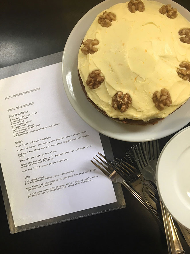 A cake decorated with orange icing and halved walnuts. A recipe and some forks lie on the table beside the cake. 