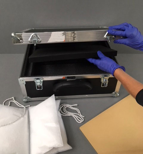 An archivist wearing nitrile gloves is closing a courier case. On the table nearby are a cushioned book rest, archive weights, and a  buff cardboard folder.