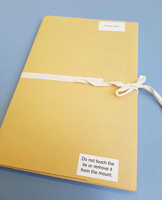 A buff-coloured folder, secured with a linen ribbon tied in a bow. There is a label on the front of the folder which reads "Do not touch the tie or remove it from its mount"