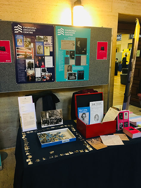 The Churchill Archives Centre stand at the History Day event. A table is arrayed with postcards, a part-completed jigsaw puzzle, and a red dispatch box which is open to reveal a variety of pamphlets etc. Behind the stall is a large board with posters about the CAC collections. 