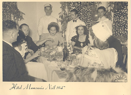 Black and white photo of a group of people including Jo Sturdee and Lady Churchill, seated at a table with waiters standing in the background. The photo is captioned 'Hotel Mamounia, Noel 1947'