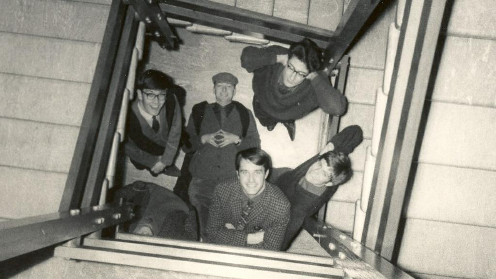Black and white photo taken from the top of a staircase looking down at a group of students standing at the bottom of the stairwell on the ground floor. One is lying on his back on the floor and all are looking up and smiling at the camera