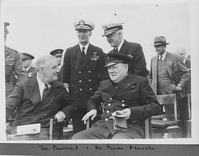 Black and white photo of FDR and Churchill seated and talking to each other, with others standing in the background. Handwritten caption reads "The President & the Prime Minister"