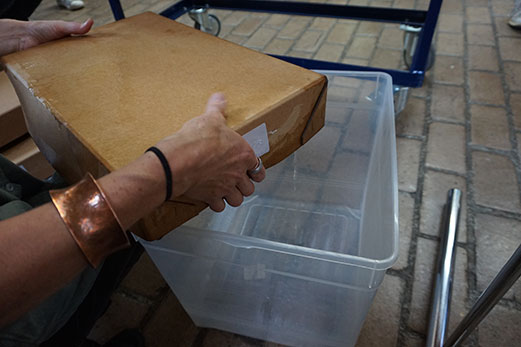 A cardboard archival box being held over a plastic crate. The cardboard box is very wet and water is draining out of it into the crate below.