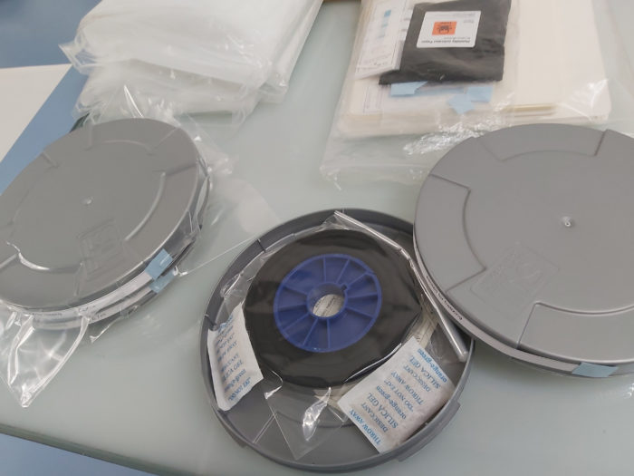 A circular metal film can lying open on a table. Inside there is a film reel inside a plastic bag, and two silica gel packets.