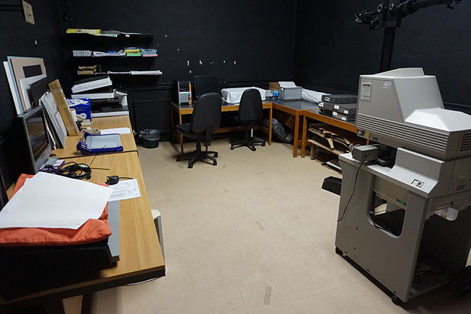 The reprographics room before the refurbishment. There are desks along three walls with a variety of computer equipment, files, and scanners.