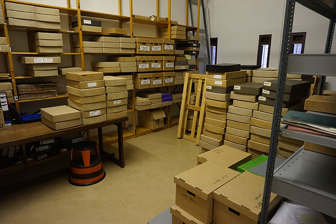The old sorting room. There are open racks of shelves on two walls, with boxes on them. A large pile of boxes is piled in between them, with more boxes on a table in the corner.
