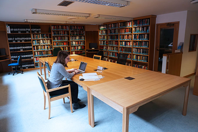 View of the reading room before the refurbishment. There is a large desk in the middle of the room with a reader sitting at it. On the far wall there are bookshelves, a computer workstation, and a door leading through to the microfilm reading room