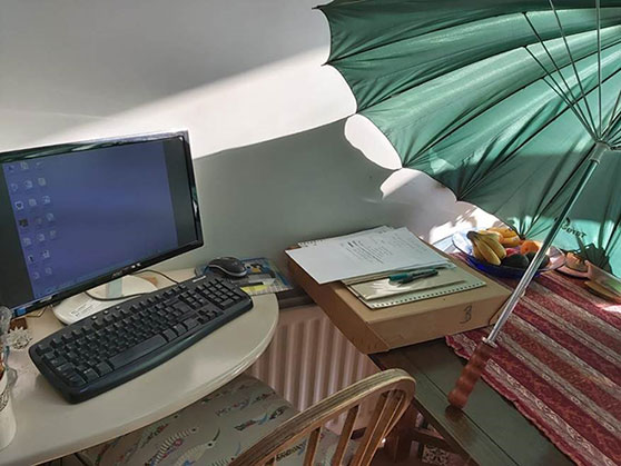 A home working set up - a desktop computer is set up on a kitchen table, a dining chair is pulled up to the table. To one side there are some files, a fruit bowl, and a large green golf umbrella which has been set up to reduce glare on the screen.