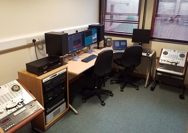 A desk with three desktop computers, a laptop, and a variety of audiovisual equipment