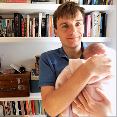 Profile photo of Tom Kelsey. He is holding a newborn baby who is wrapped in a pink blanket, and they are standing in front of a bookcase