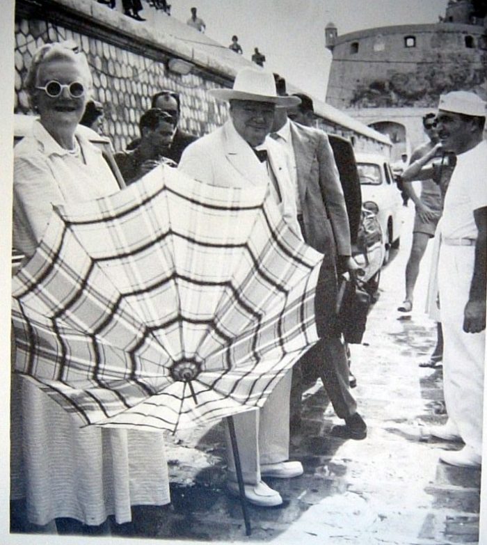 Black and white photo of Clementine and Winston Churchill. Clementine is wearing a white dress, sunglasses and holding a parasol, Winston is wearing a white suit and hat.