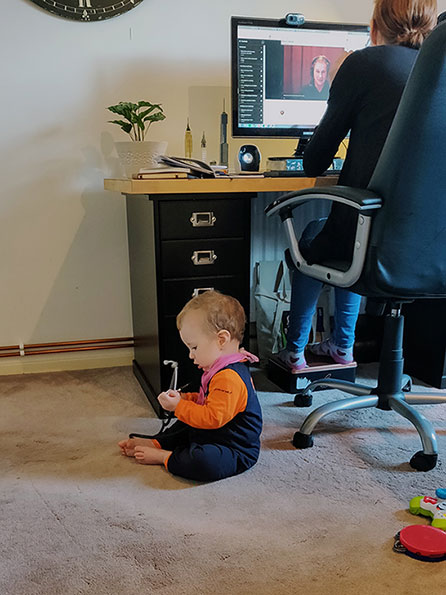 A woman sits in an office chair at a desk taking part in an online meeting on her computer. At her feet a toddler sits on the floor playing with a stethoscope.