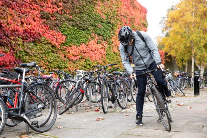 Male student setting off on a bike from the front of College. In the background are a number of bike racks and the Boston Ivy on the walls of the Squash Courts.