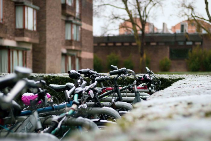 A row of bicycles in the snow, with College buildings in the background. A light dusting of snow sits on the bikes and on the surrounding hedge.