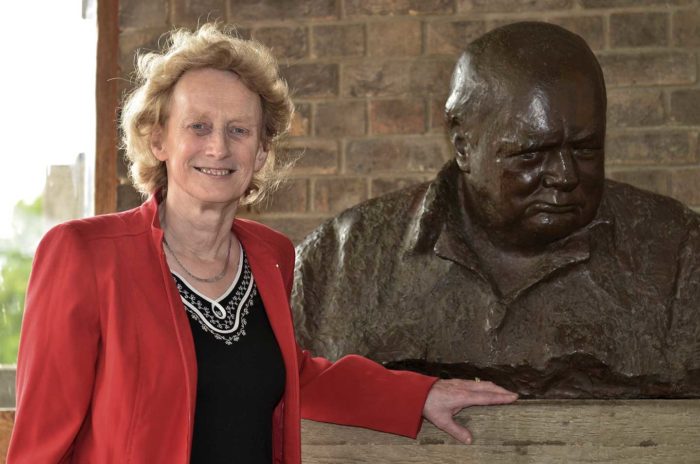 Dame Athene Donald standing next to a bust of Churchill in College. Her hand is resting on the plinth and she is smiling at the camera.
