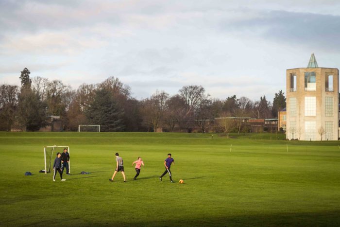 Five students play football on the College playing field. The Moller Centre tower (a hexagonal white building with a glass spire) can be seen at the right hand side of the photo