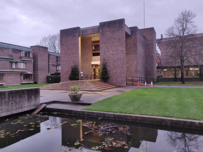 The front of College at sunrise. There are Christmas trees on either side of the entrance, and the building is reflected in the pond.
