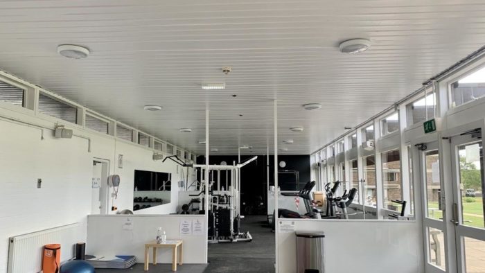 Interior shot of the College gym. In the foreground there is a yoga mat and exercise ball, further down the gym there is a variety of equipment including treadmills, rowing machines and squat racks.