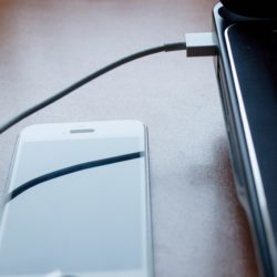 A smartphone lying on a desk next to a laptop, connected by a USB charging cable