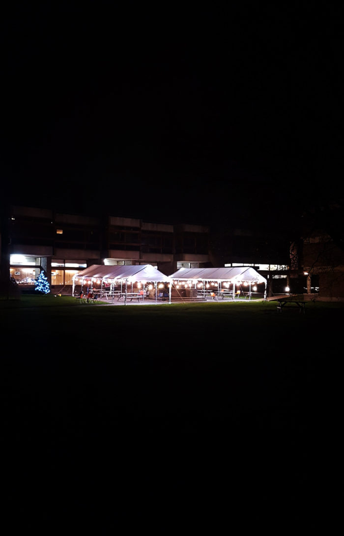 A night time shot of two marquees, with the main concourse building in the background. There are lights on the marquees, and a lit Christmas tree can be seen beside them.