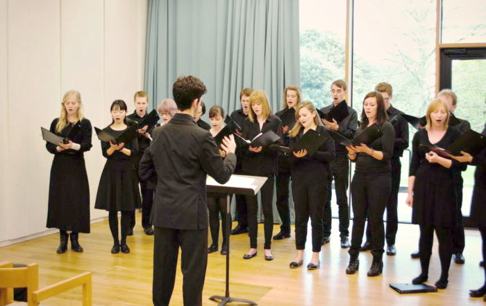 A conductor rehearsing with the choir, who are dressed in black and holding music folders.