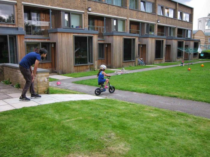 A young girl rides a bike in the play area outside the Wolfson Flats, with her father standing behind her.