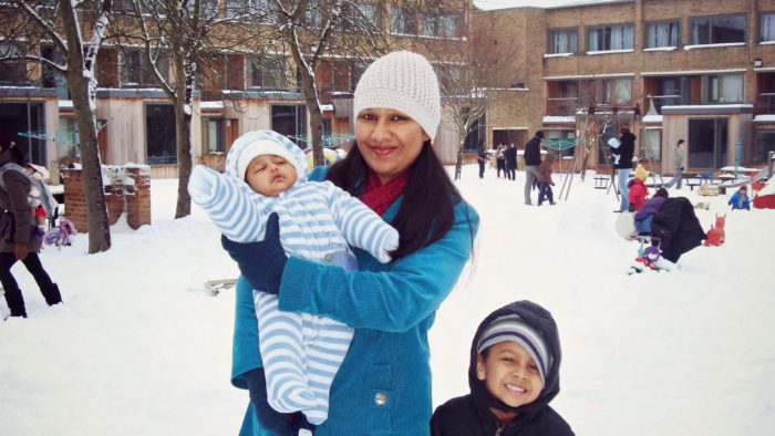 A female postgraduate student and her family standing in the snow smiling at the camera. In the playground behind her lots of families are playing in the snow.