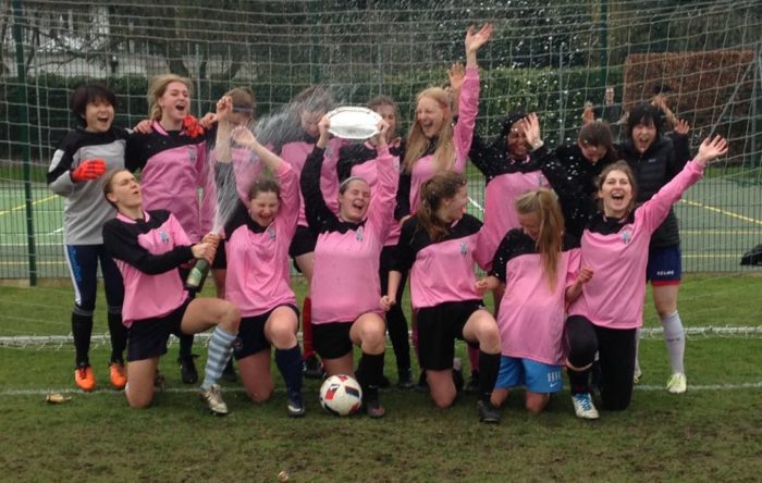 The women's football Cuppers team celebrating a victory in 2018. A player in the centre holds a silver plate while another player sprays champagne over the team