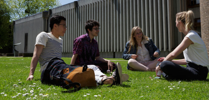 A group of four students sitting on the grass outside, with the Library and Archives Centre in the background