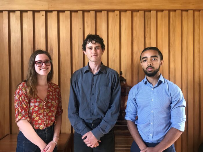 One female and two male students standing in front of a panelled wood wall. The female student is wearing a red top, and the male students are wearing blue shirts.