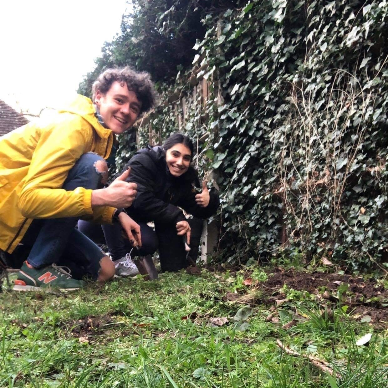 Two students kneeling in the garden, smiling and giving a thumbs up to the camera
