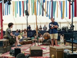 An image of two performers at the Wilderness Music Festival performing 'Hebrides Redacted' in a tented arena. Multicoloured streamers are hanging above the performance space which is furnished with travelling trunks and chairs.