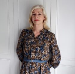 Colour half length photo of Sharon Mather, wearing a blue and brown dress and leaning back against white doors