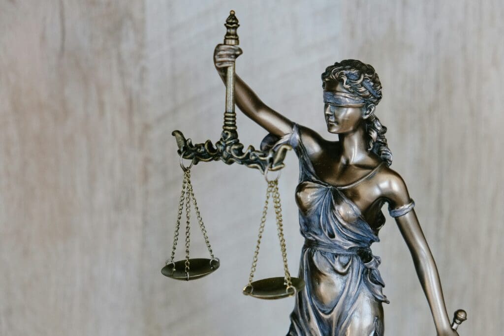 A bronze statue of Lady Justice, representing the law, is shown holding a balanced scale in her right hand and a sword in her left. She is blindfolded, symbolizing impartiality, with her figure draped in classical robes against a neutral background.