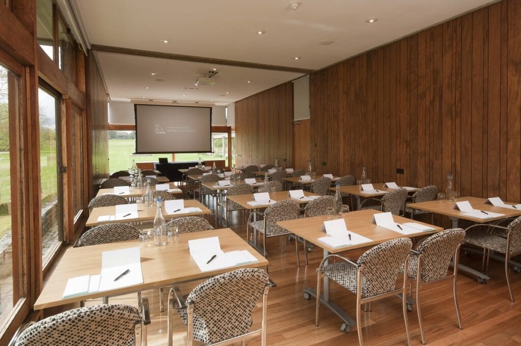 A conference room with wooden walls and floor, featuring several rows of tables and chairs. Each table has writing materials and water bottles. A large screen at the front displays a presentation. Natural light enters through large windows on the left.
