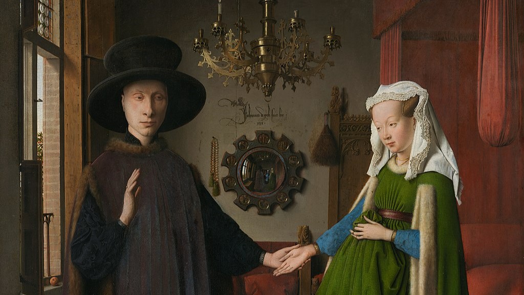 A man and woman stand holding hands in a 15th-century room. The man wears a dark robe and hat, while the woman wears a green dress with a white headdress. A chandelier hangs above them, and a mirror and bed are in the background.