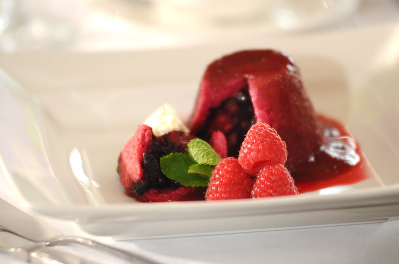 A dessert with a red, dome-shaped cake, featuring a glossy berry sauce. It is garnished with fresh raspberries, mint leaves, and a dollop of cream, presented on a white plate.
