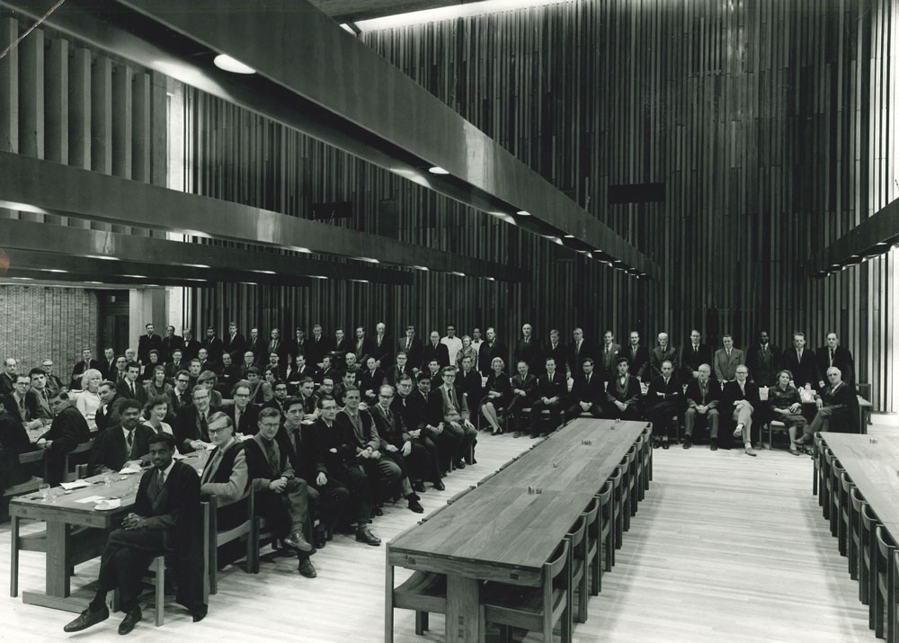 A large group of people sits and stands in a spacious hall with wooden beams and long tables. They are arranged to face the camera, dressed in formal to semi-formal attire. The setting has a modern architectural style.