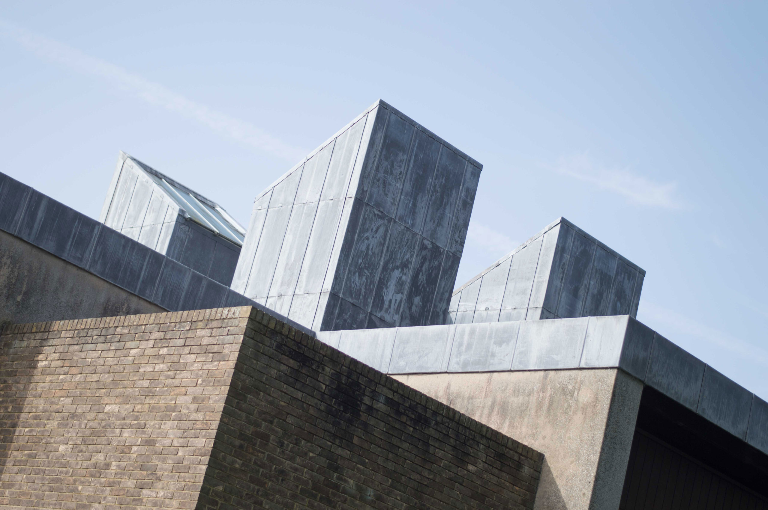 Modern architectural structure with uneven, geometric metal and brick elements against a clear blue sky. The design features angular metal rooftops and textured brick walls, creating a contemporary aesthetic.