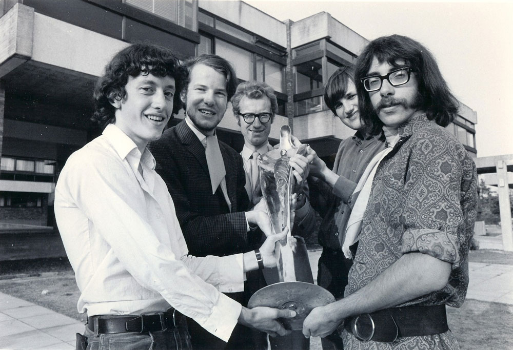 Five men stand outdoors in front of a building. They are smiling and holding a trophy from University Challenge.